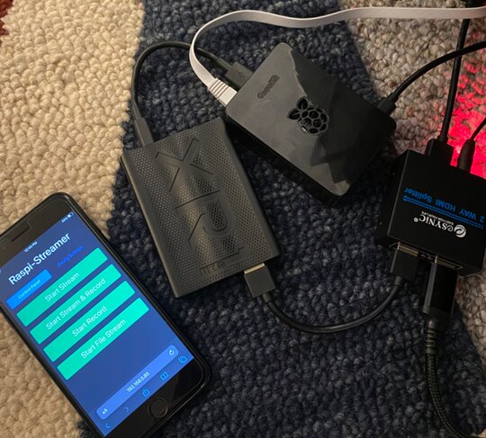 Cellphone with the Raspi-Streamer web UI open, a USB caputer device connected to a Raspberry Pi 5, a HDMI splitter with one hdmi cable going to the capture device and another cable going to the tv.